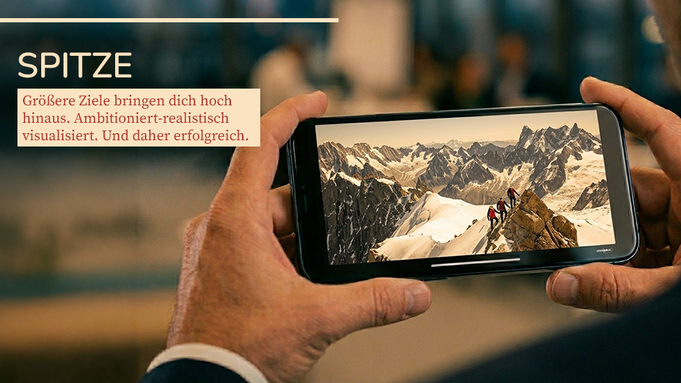 Unternehmer im Großraumbüro hält ein Smartphone waagerecht in seinen Händen. Darauf sind ein Bergpanorama und eine aufsteigende Gruppe zu sehen. Headline: Spitze. Text darunter: Größere Ziele bringen dich hoch hinaus. Ambitioniert-realistisch visualisiert. Und daher erfolgreich.